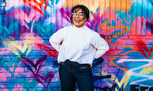 Woman with glasses standing in front of colorful wall.